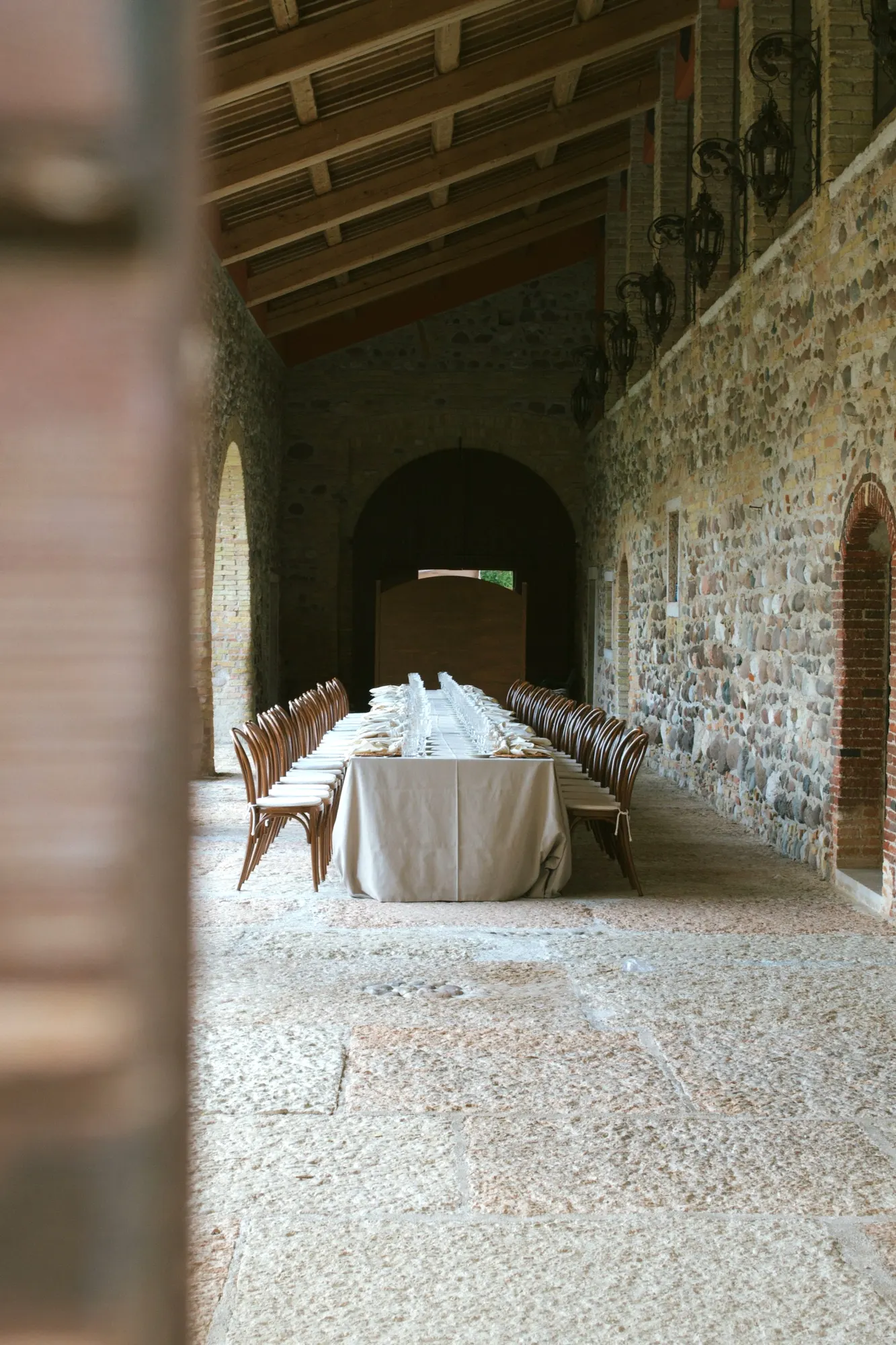 Long banquet table laid in a stone loggia with a timber ceiling